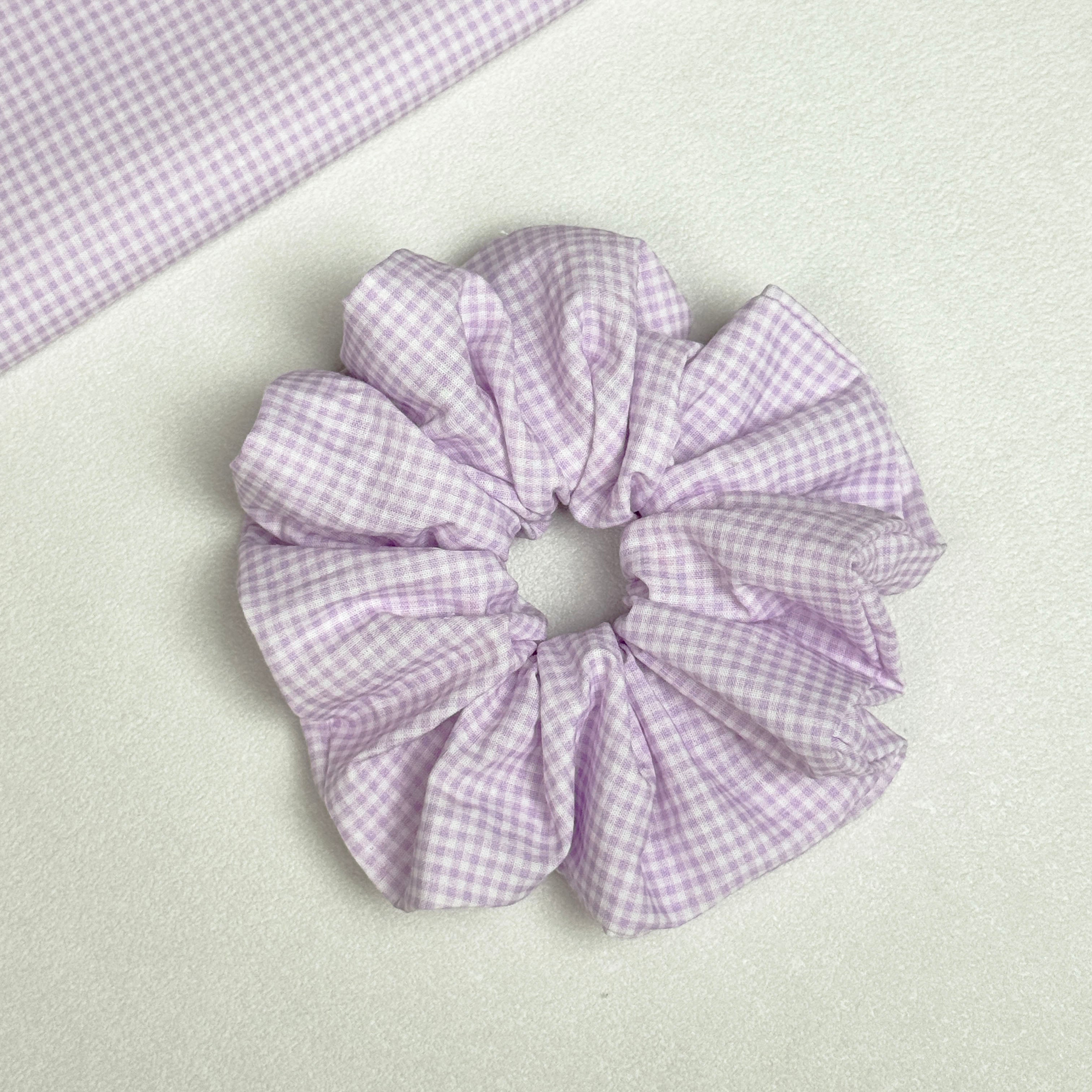Lavender gingham scrunchie displayed alongside a matching fabric swatch laid diagonally, showing the full gingham check pattern, white pebbled texture surface.