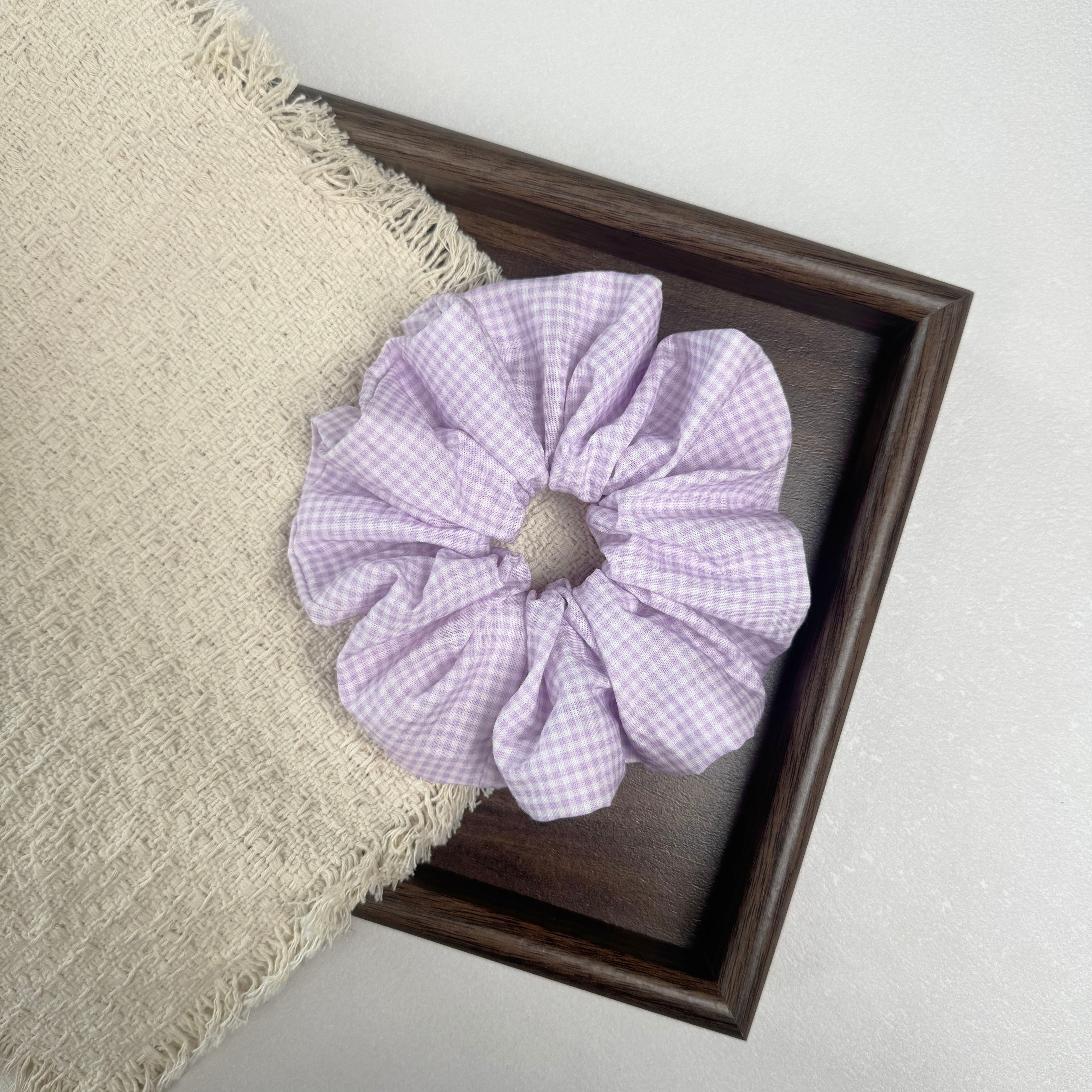 Lavender gingham scrunchie flat-laid across a dark brown wooden tray and a frayed cream woven fabric with tasseled edges, grey-white textured surface.