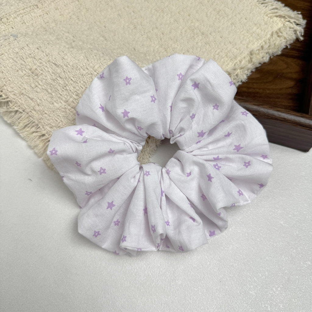 White lavender star print scrunchie on a grey-white surface beside a frayed cream burlap-textured fabric and the corner of a dark brown wooden tray, flat-lay composition.