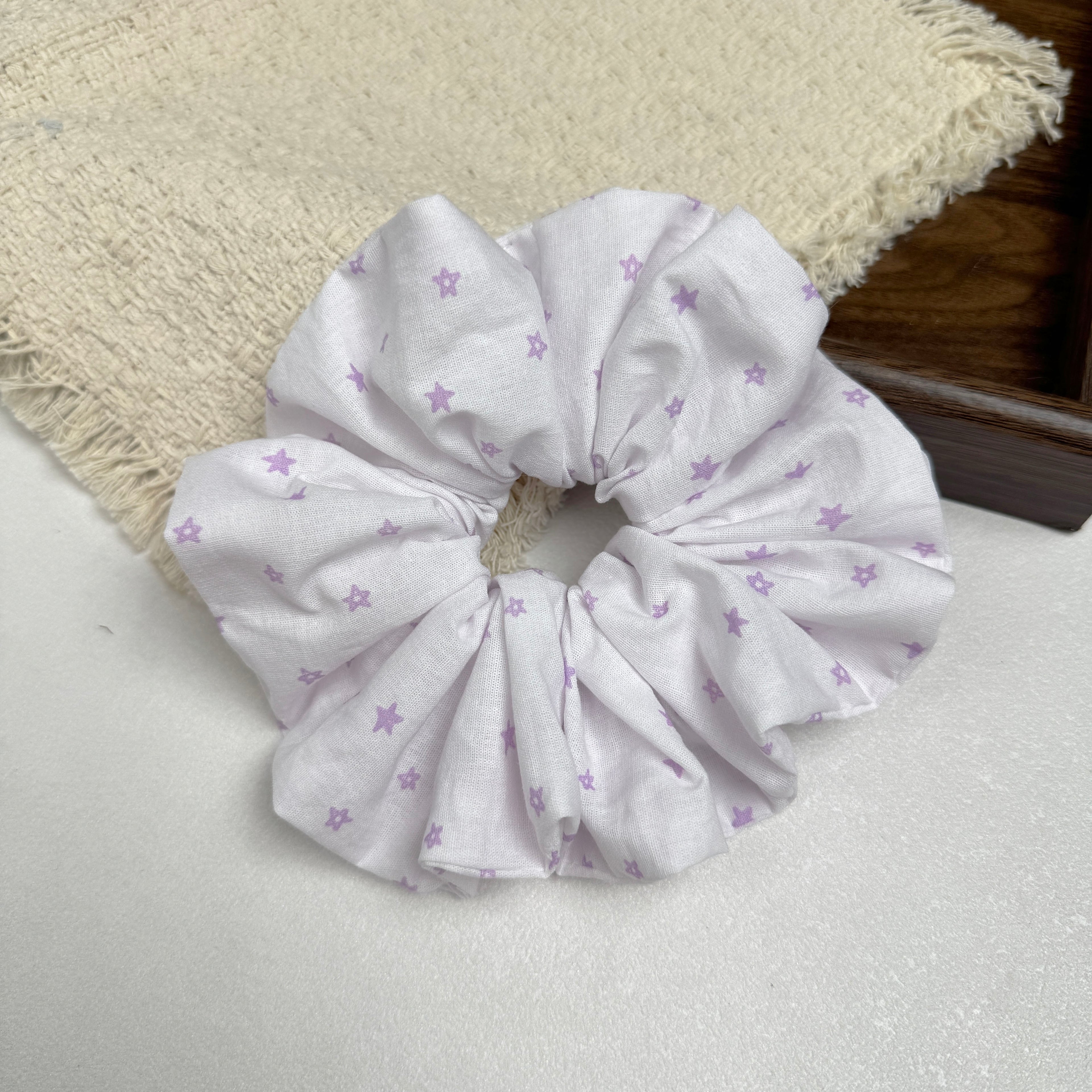 White lavender star print scrunchie on a grey-white surface beside a frayed cream burlap-textured fabric and the corner of a dark brown wooden tray, flat-lay composition.