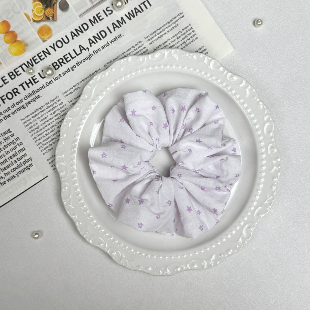 White lavender star print scrunchie placed on a white ceramic plate with embossed scalloped rim, resting on a magazine page with citrus photography and text, scattered white faux pearls, grey-white surface.
