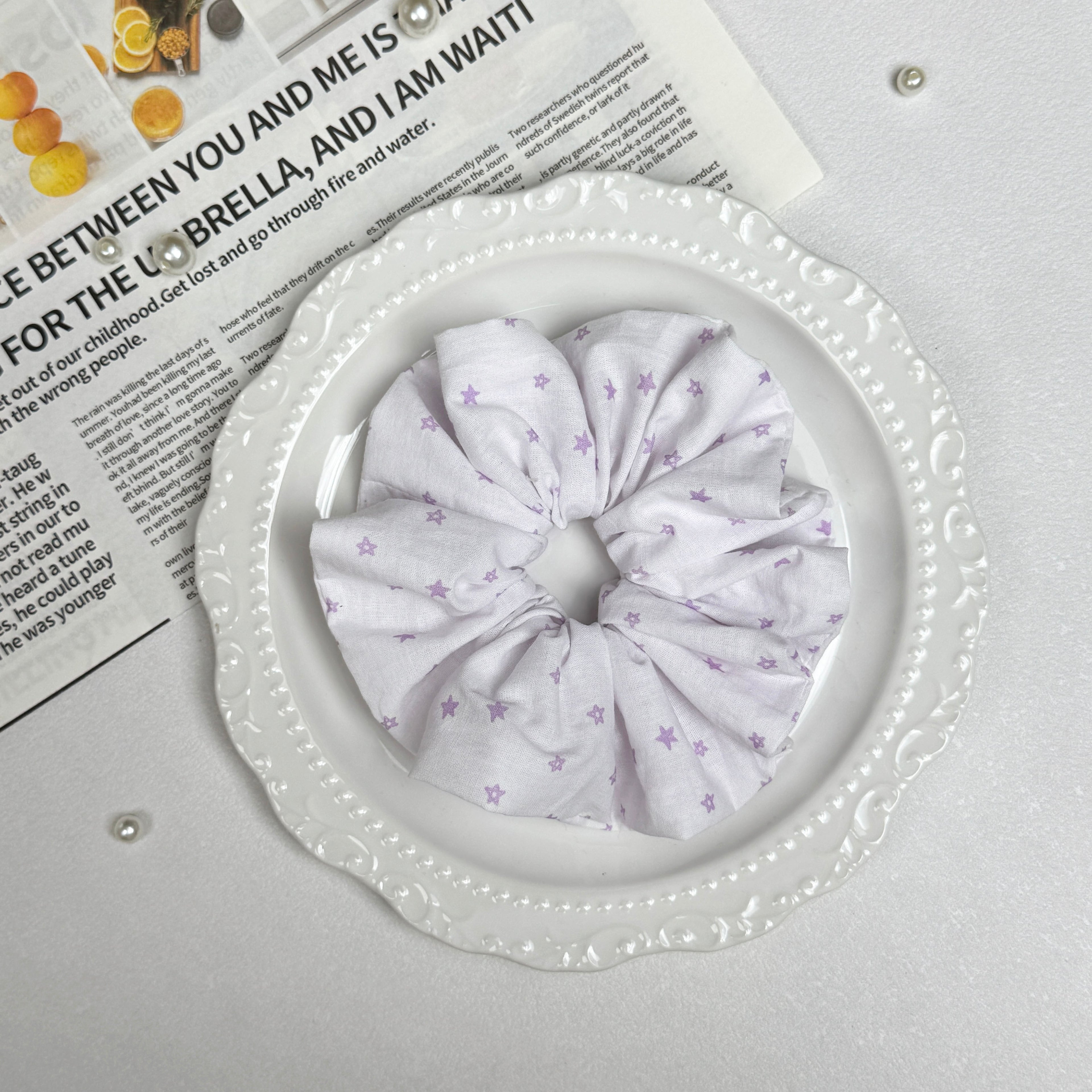 White lavender star print scrunchie placed on a white ceramic plate with embossed scalloped rim, resting on a magazine page with citrus photography and text, scattered white faux pearls, grey-white surface.