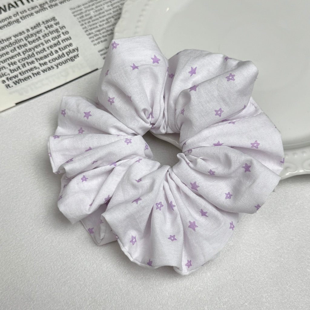 White lavender star print scrunchie alongside a white ceramic plate with beaded scalloped rim and an open magazine page with printed text, clean white surface, flat-lay composition.