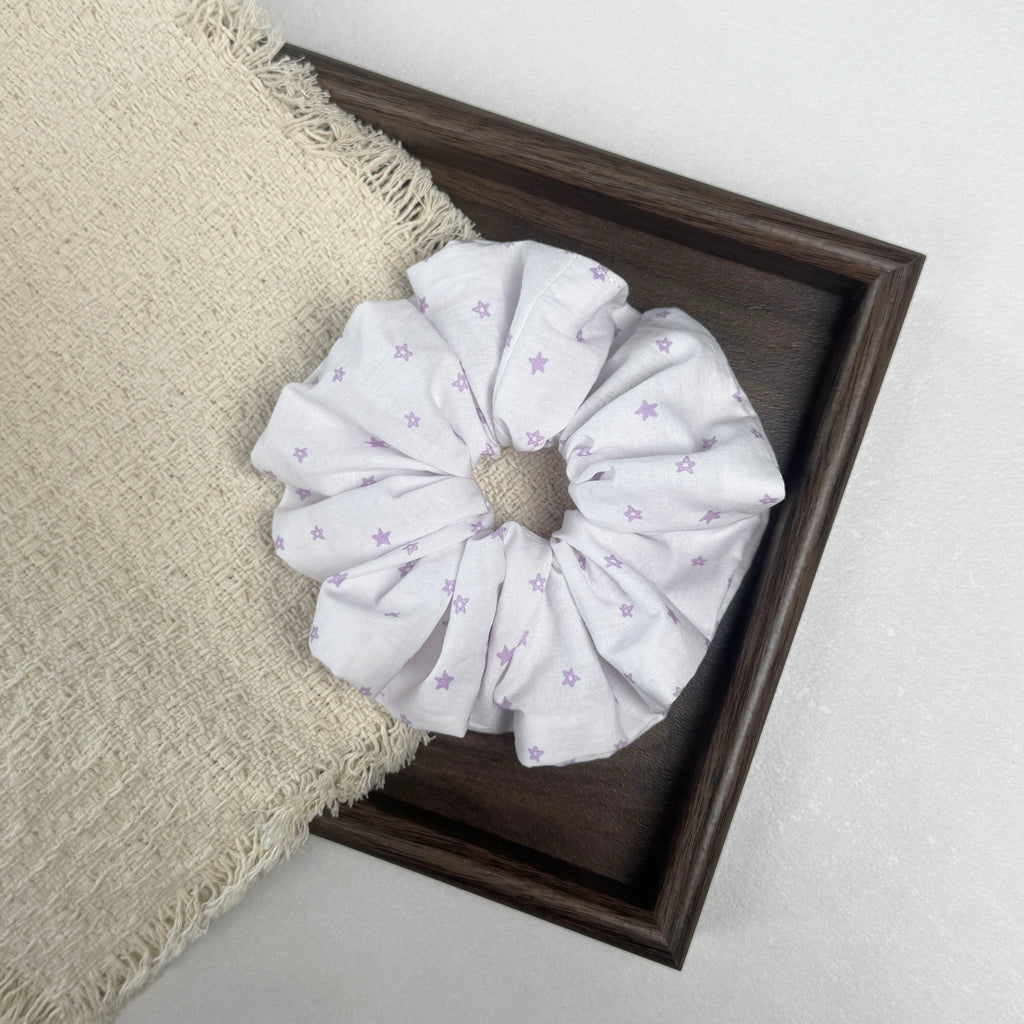 White lavender star print scrunchie flat-laid across a dark brown wooden tray and a frayed cream waffle-weave fabric with tasseled edges, grey-white textured surface.