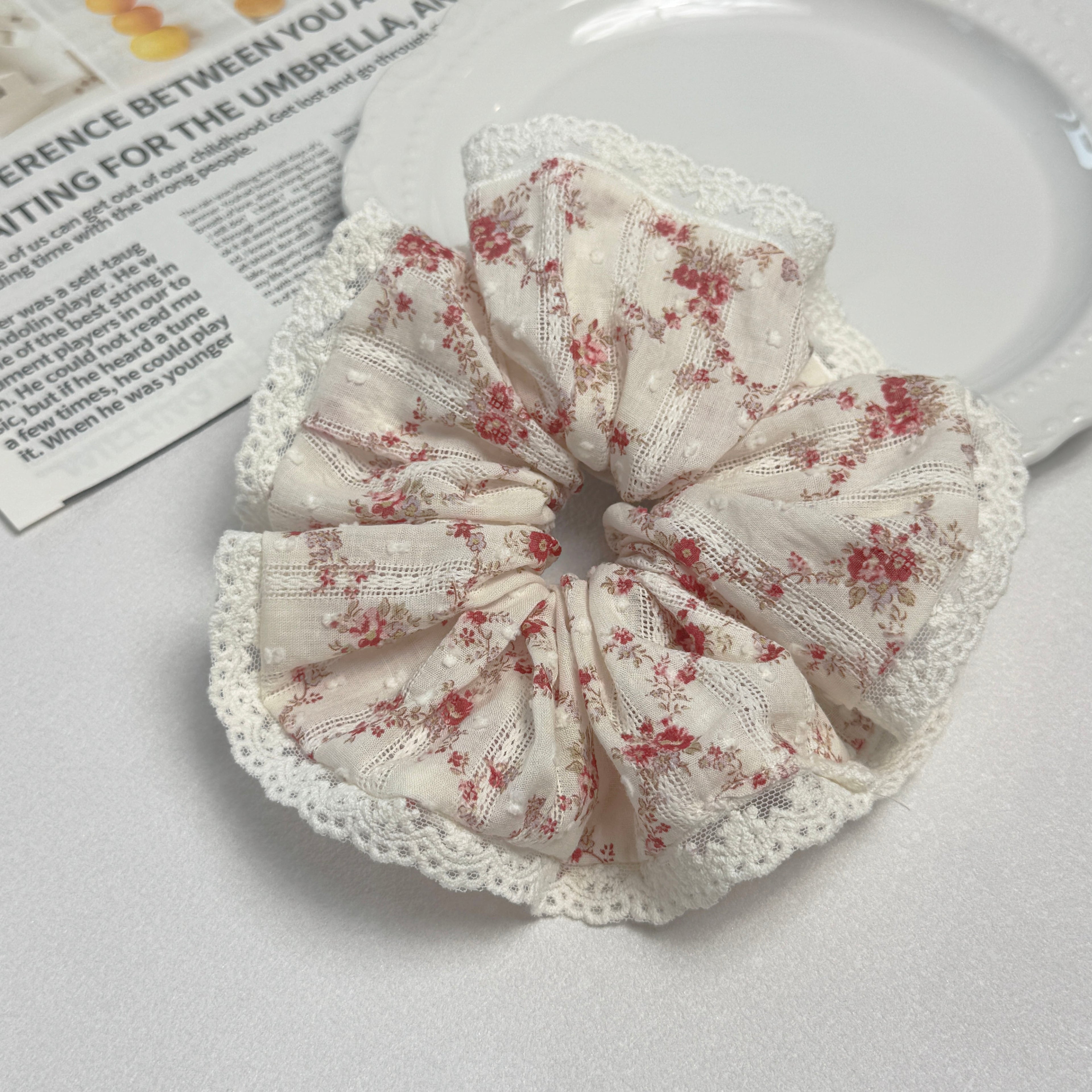 Red floral lace scrunchie on a white textured surface beside a white beaded-rim plate and a magazine page with fruit photography, top-down flat lay.