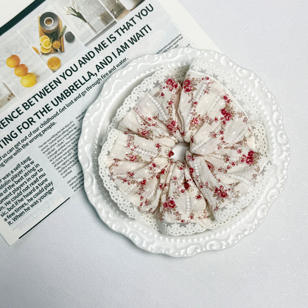 Red floral lace scrunchie placed on a white ceramic scalloped plate, resting on an open magazine page with small food photography, white surface.