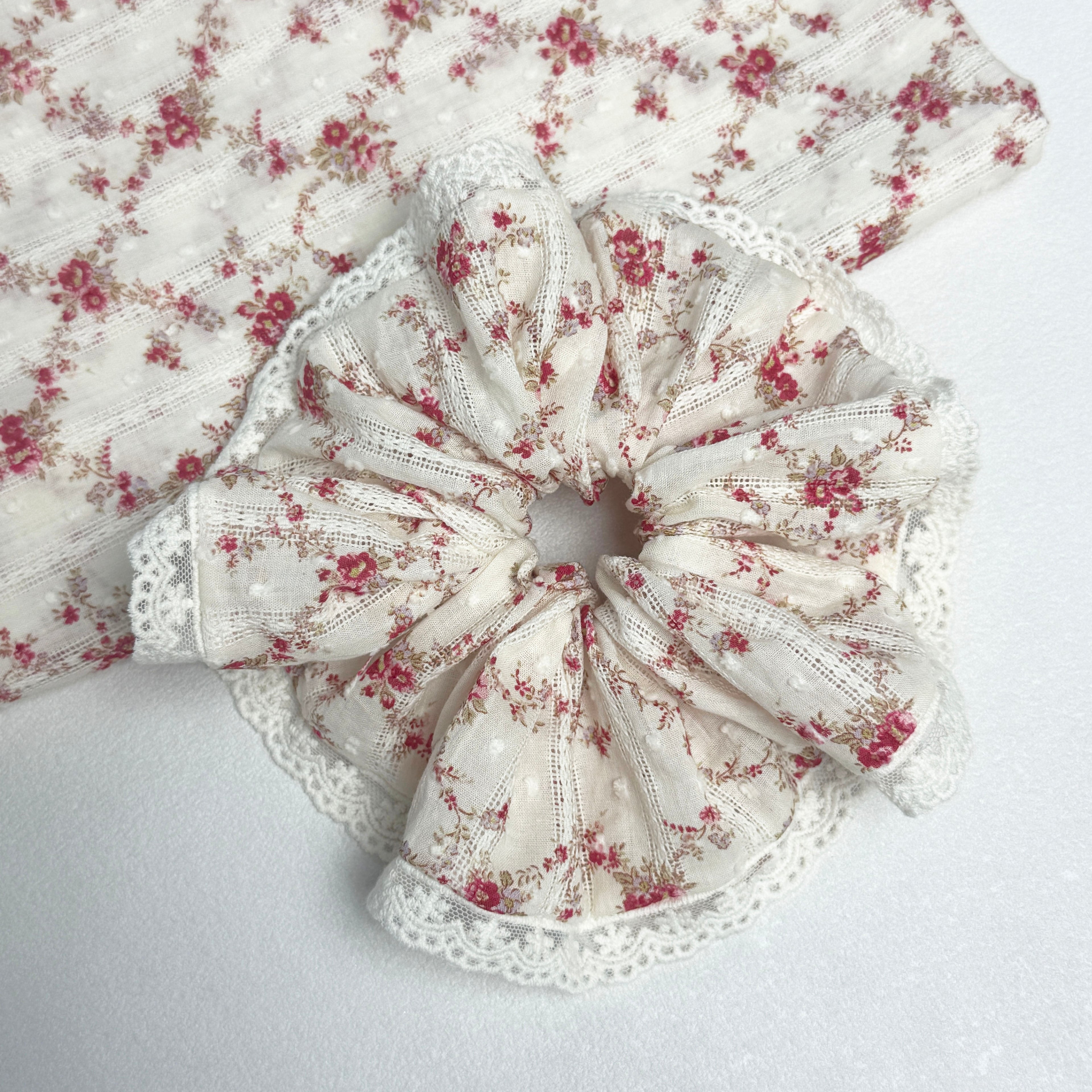 Red floral lace scrunchie displayed alongside a matching folded fabric swatch showing the floral print and vertical eyelet stripes, white textured surface.