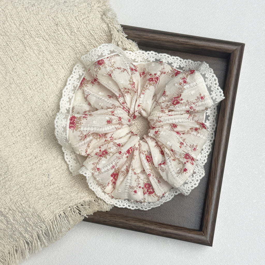 Red floral lace scrunchie flat-laid across a dark brown wooden frame and a frayed cream woven fabric, white textured surface.