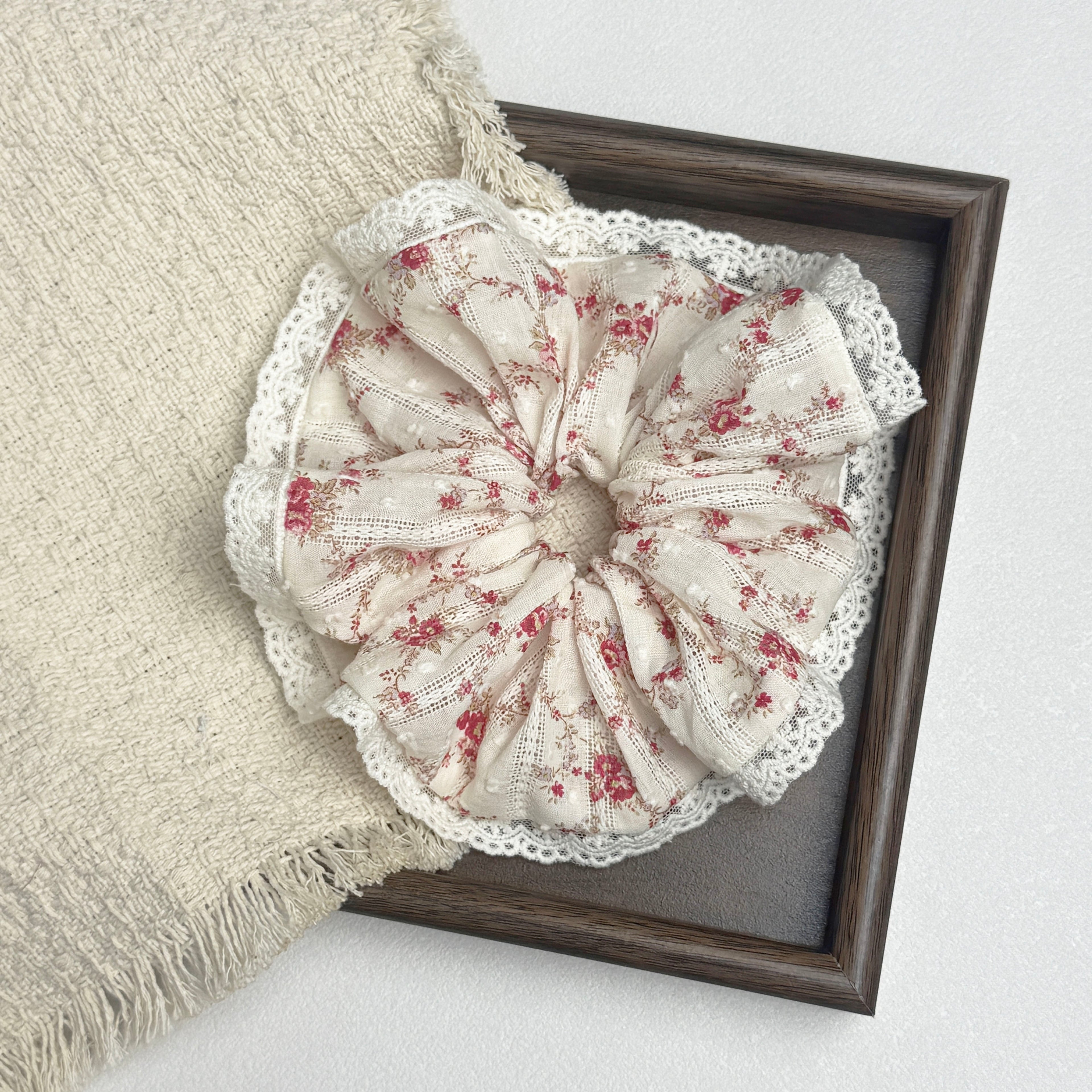 Red floral lace scrunchie flat-laid across a dark brown wooden frame and a frayed cream woven fabric, white textured surface.