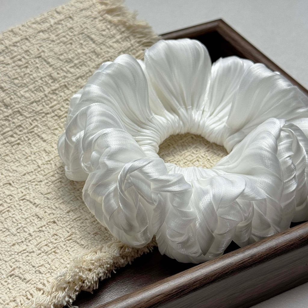 White organza cloud scrunchie on a cream bouclé textured cloth with frayed edges, dark brown wooden frame edges visible at right and bottom, grey-white surface.