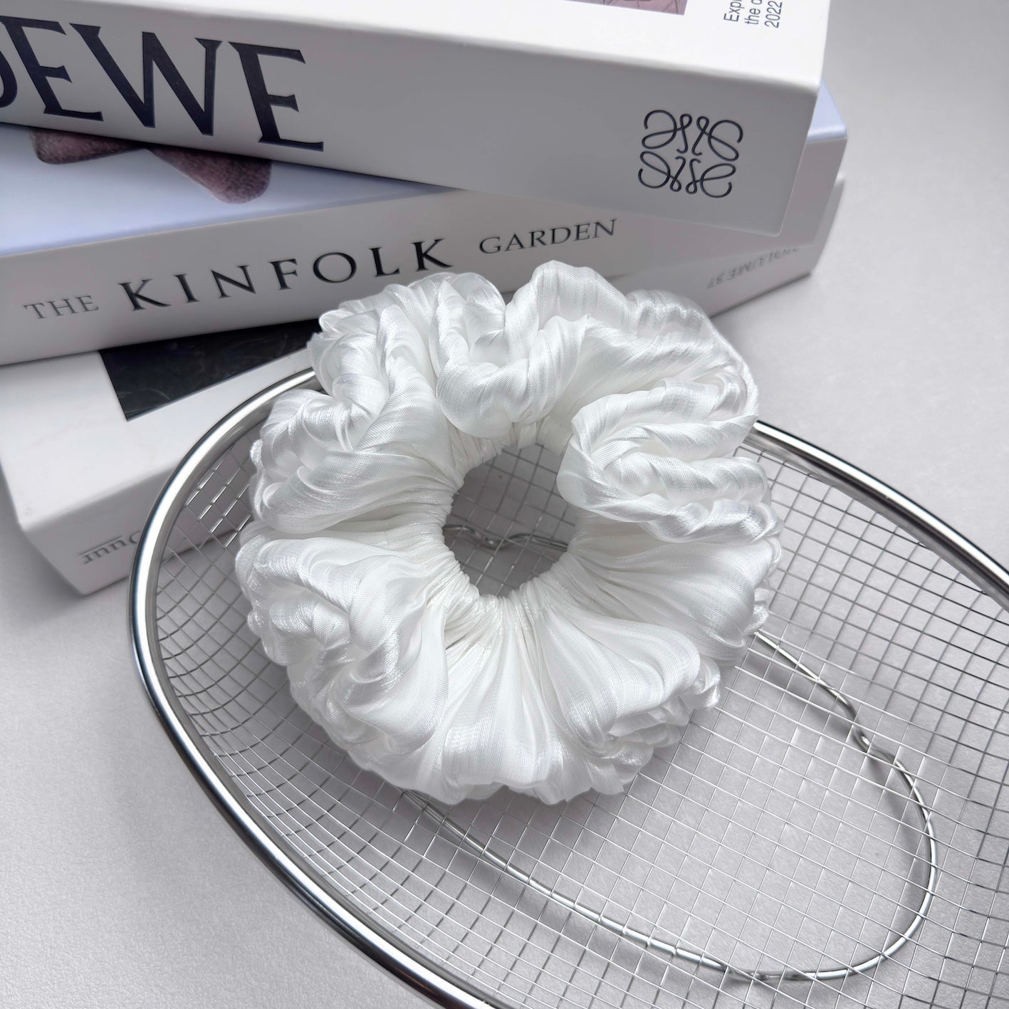 White organza cloud scrunchie resting in a polished silver wire mesh basket, placed on stacked white books, light grey surface.