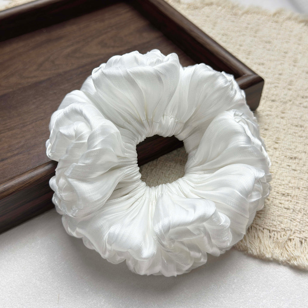 White organza cloud scrunchie resting on a fringed cream woven textile, with the corner of a dark brown wooden tray visible, grey-white surface, flat-lay composition.