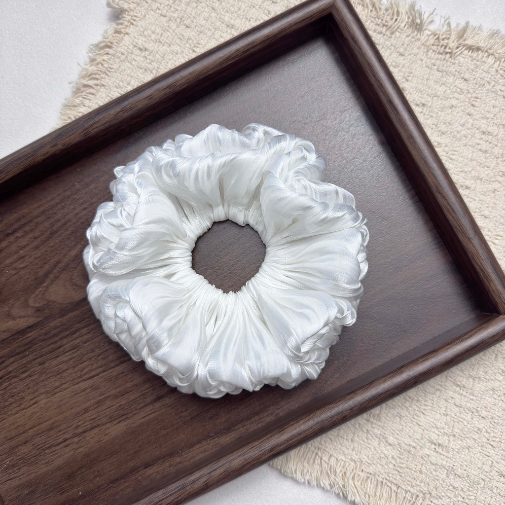 White organza cloud scrunchie centered in a dark brown wooden tray with prominent grain, resting on a fringed cream woven textile, grey-white surface, flat-lay composition.