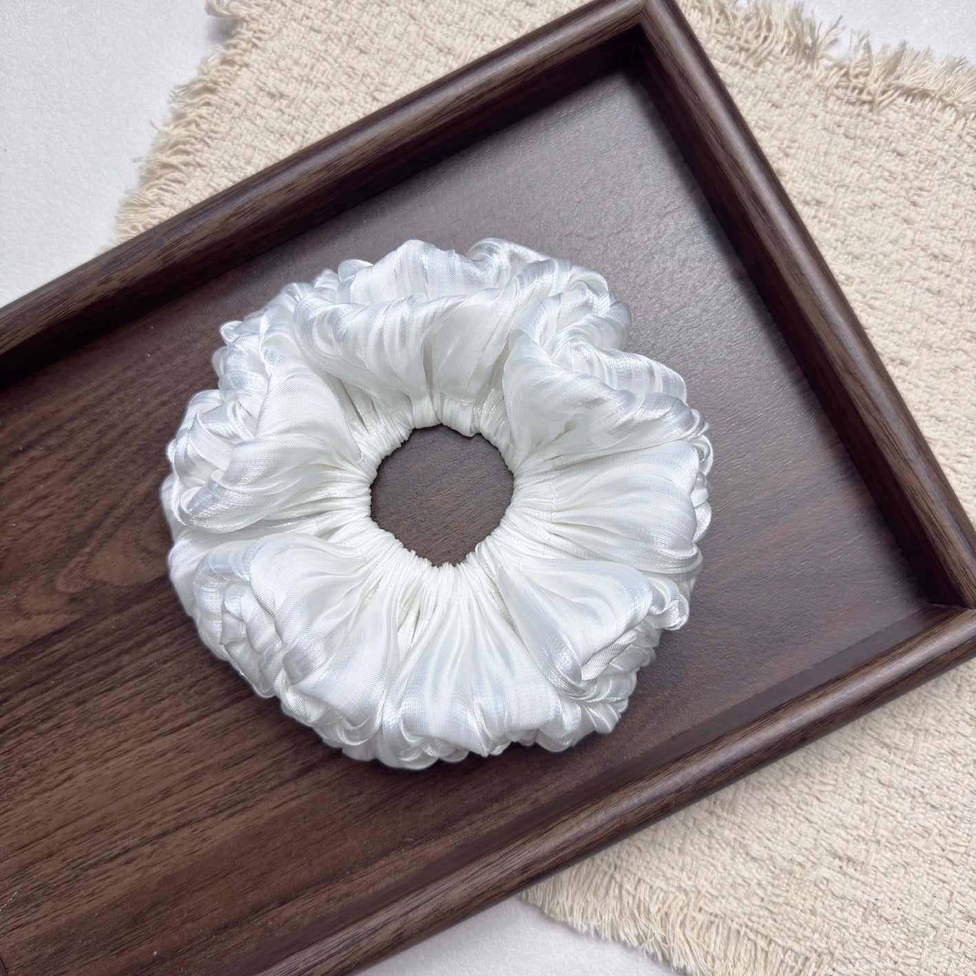 White organza cloud scrunchie centered in a dark brown wooden tray with prominent grain, resting on a fringed cream woven textile, grey-white surface, flat-lay composition.