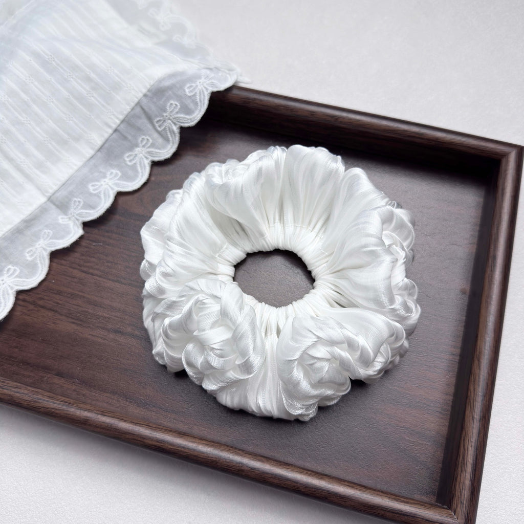 White organza cloud scrunchie in a dark brown wooden tray, a piece of white semi-sheer fabric with pinstripes and floral lace trim overlapping the top left corner, grey-white surface.