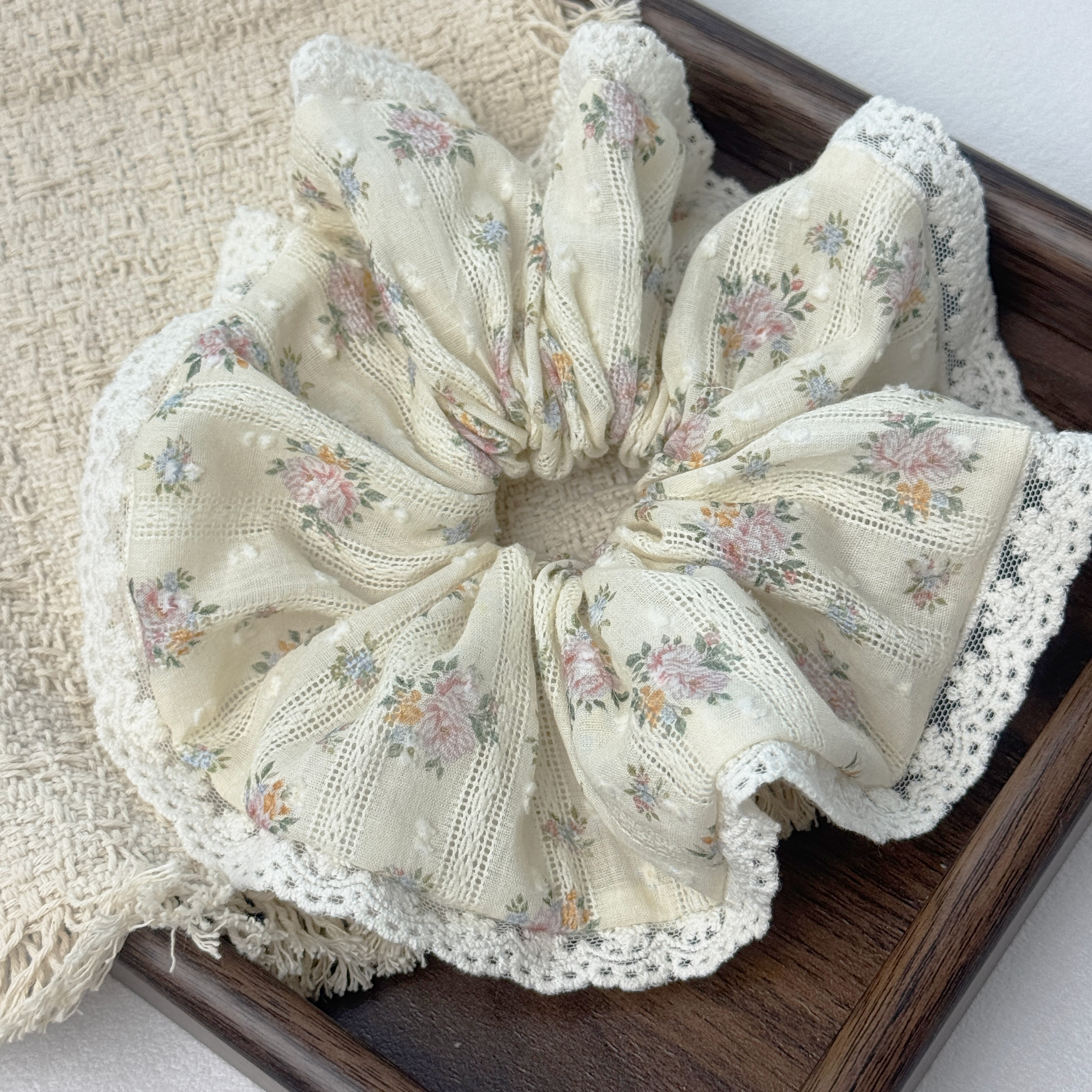 Close-up top view of a yellow floral lace scrunchie on a wooden tray, showcasing lace trim and linen pleats.