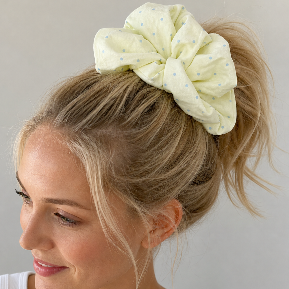 Blonde woman wearing a pastel yellow polka dot scrunchie in a high ponytail, side profile against a neutral off-white background.