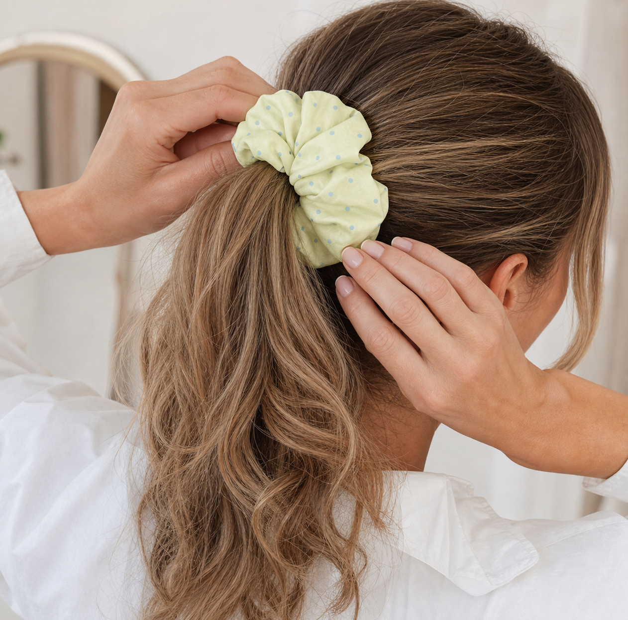 Hands pulling a pastel yellow polka dot scrunchie over blonde wavy hair into a ponytail, rear view. Mirror in blurred indoor background.