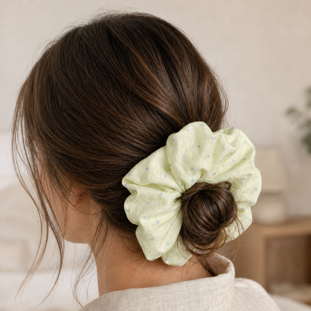 Pastel yellow polka dot scrunchie styled in a low bun, rear view. Soft-focus domestic interior background.