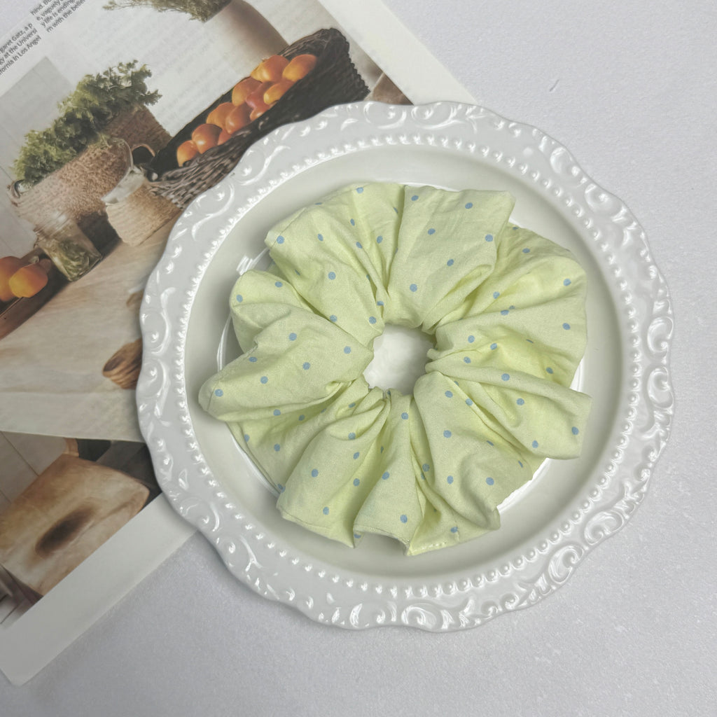 Pastel yellow polka dot scrunchie placed in a white ceramic scalloped plate, resting on an open magazine page, white surface.