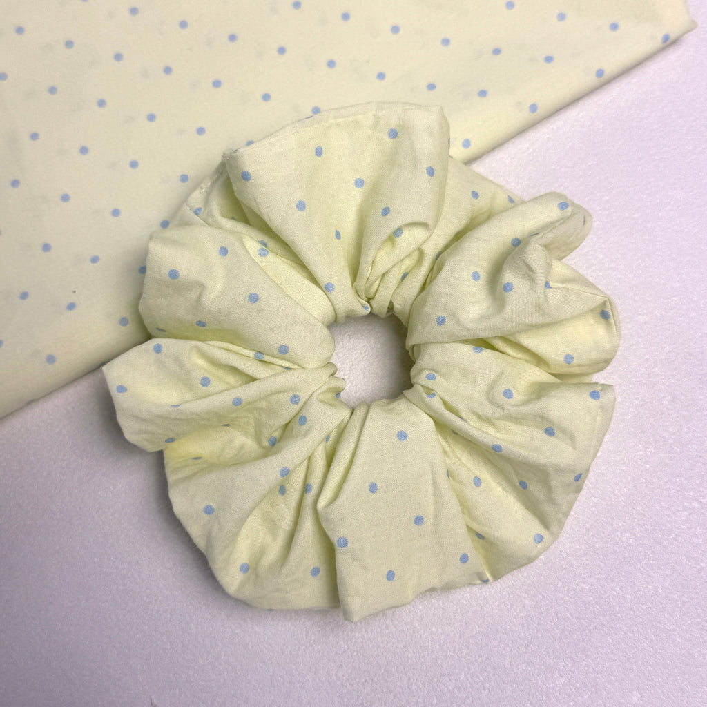 Pastel yellow polka dot scrunchie displayed alongside a matching fabric swatch on a white textured surface.
