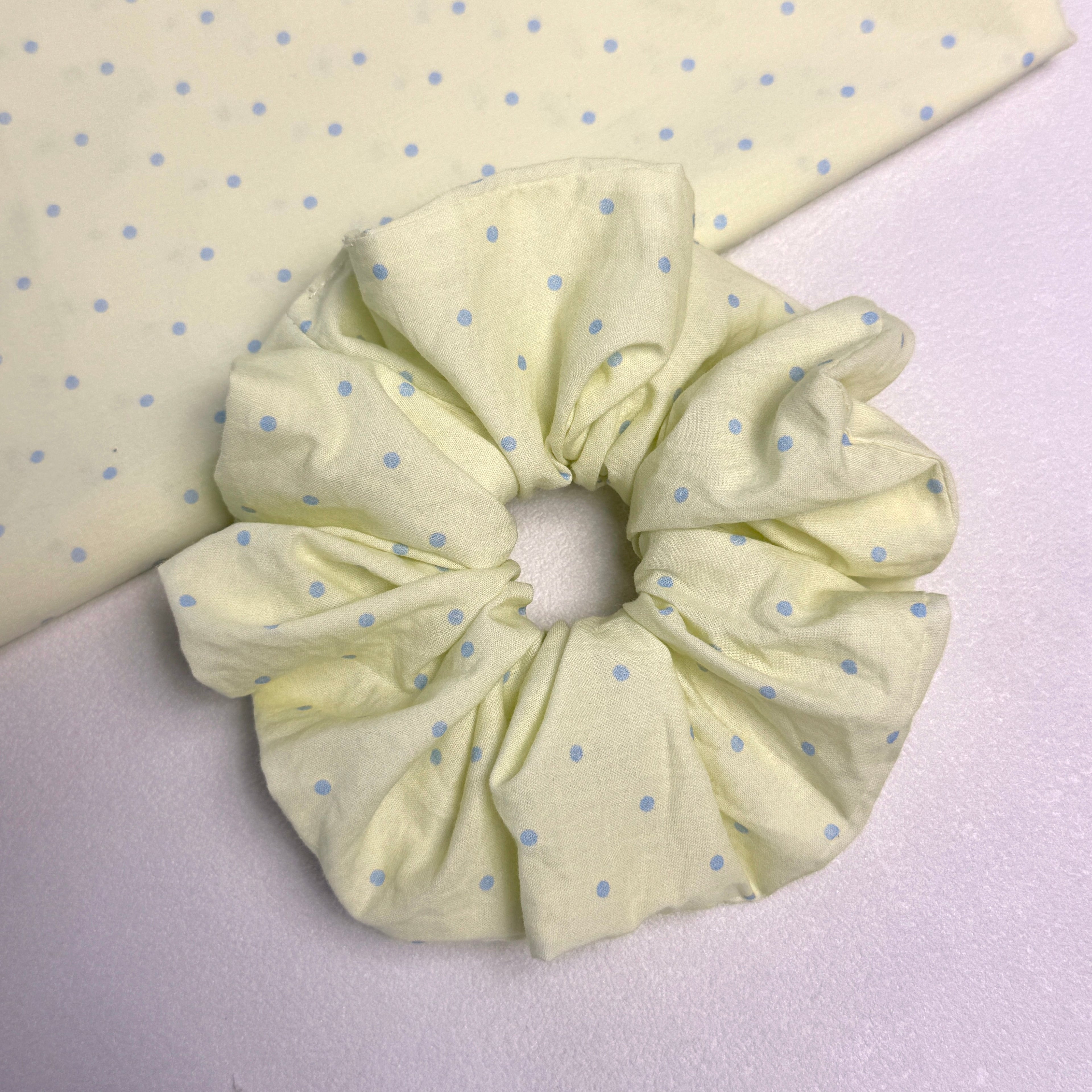 Pastel yellow polka dot scrunchie displayed alongside a matching fabric swatch on a white textured surface.
