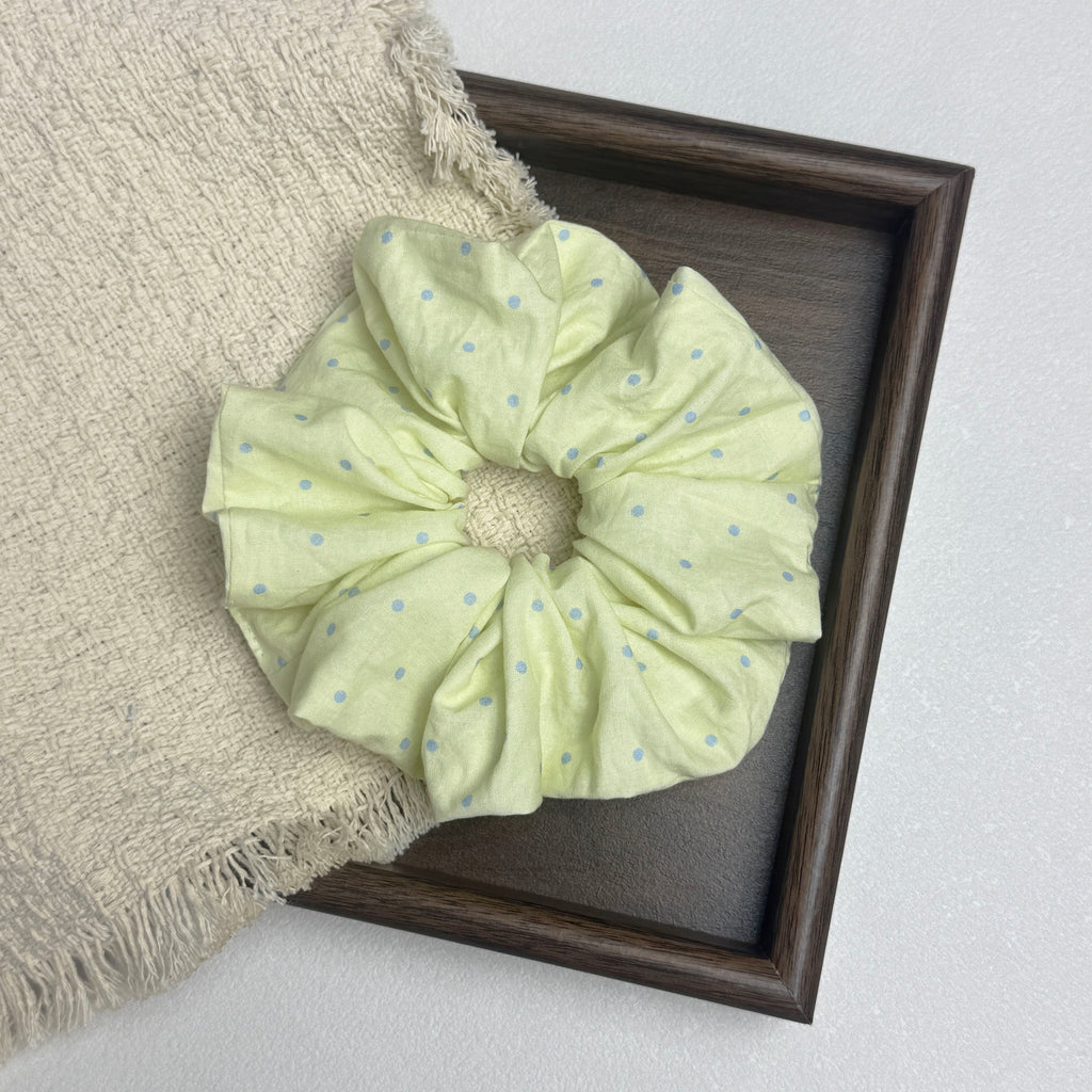 Pastel yellow polka dot scrunchie flat-laid across a dark wood tray and an off-white fringed woven cloth, white surface.