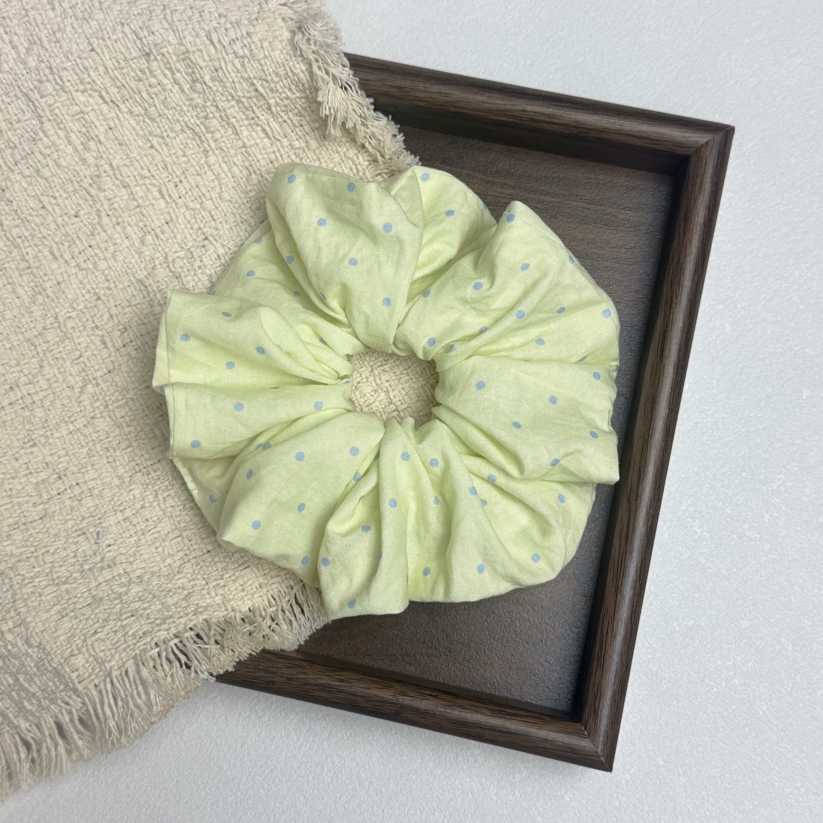 Pastel yellow polka dot scrunchie flat-laid across a dark wood tray and an off-white fringed woven cloth, white surface.